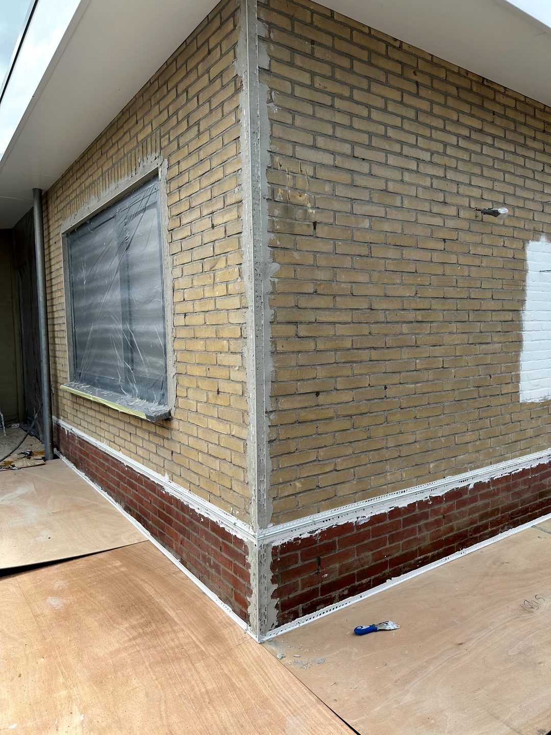Bakstenen hoek van de bungalow tijdens de voorbereiding: kozijnen afgeplakt en hoek- en plintprofielen geplaatst